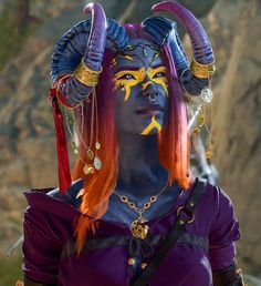 A blue tiefling woman with purple and orange hair, wearing yellow and purple face paint, stands with many thin chains and pendants hanging from her horns. She smiles, while looking upwards. Baldurs Gate 3 Custom Characters, Bg3 Githyanki, Bg3 Character Creation No Mods, Bg3 Bard