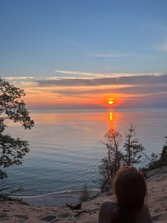 a woman sitting on top of a sandy hill next to the ocean at sunset or sunrise