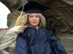 a doll wearing a graduation cap and gown with long blonde hair in front of a rock formation