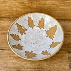 a white and gold bowl with trees painted on the side sitting on a wooden floor