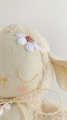 a white stuffed animal with a flower on it's head and eyes, sitting in front of a white wall