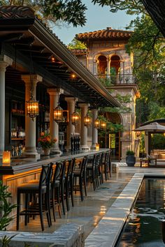 an outdoor bar with tables and chairs next to a pool at dusk, surrounded by greenery