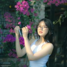 a woman in white shirt and glasses holding up cell phone next to purple flower bush