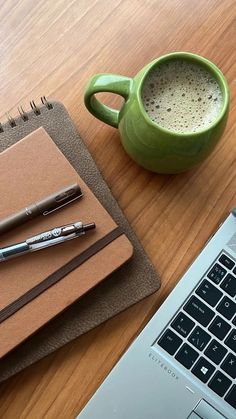 a cup of coffee next to a notebook and pen on a wooden table with a laptop