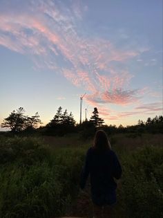 a woman walking through tall grass towards a wind turbine in the distance with pink and blue clouds