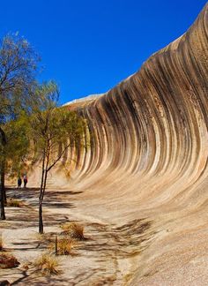 two people are walking on the side of a large rock formation with trees in front of it