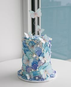 there is a blue and white cake with butterflies on the top, sitting on a table in front of a window