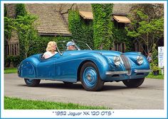 an old blue sports car with a woman in the driver's seat driving past a house