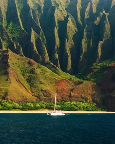 a sailboat in the water near some green mountains and sand beach with trees on both sides