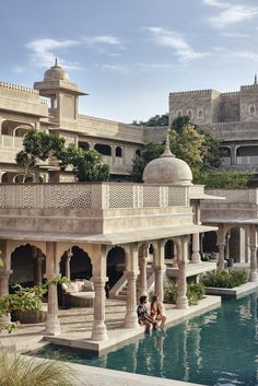 two people sitting on the edge of a pool in front of a palace like building