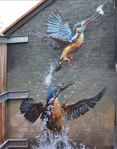 two birds are flying over the water in front of a mural on a brick wall