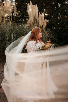 a woman wearing a wedding dress and veil standing in front of some tall grass with her hair blowing in the wind