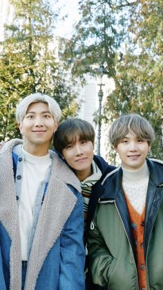 three young men standing next to each other in front of some trees and buildings with one man wearing a blue jacket