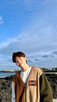 a young man standing next to the ocean with his hands in his pockets and looking at the camera