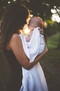 a woman holding a baby wrapped in a white towel with the sun shining through trees behind her