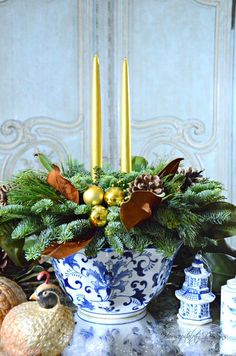 a blue and white bowl filled with greenery next to two gold candlesticks