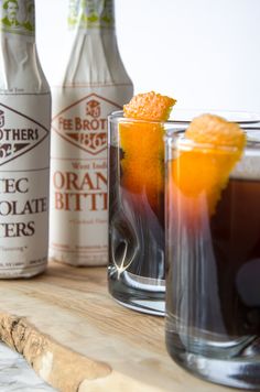 two glasses filled with liquid and an orange slice next to bottles on a wooden table