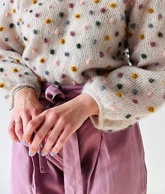 a woman with her hands on her hip wearing purple pants and a white sweater that has multicolored polka dots