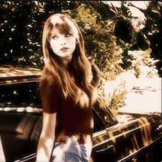 a woman standing next to a parked car in front of some trees and bushes with her hand on her hip