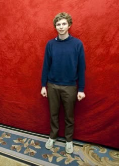 a young man standing in front of a red wall with his hands in his pockets