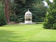 a white gazebo in the middle of a green lawn surrounded by trees and bushes