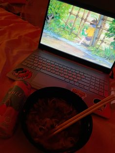 an open laptop computer sitting on top of a bed next to a bowl of noodles