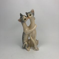 two ceramic cats sitting next to each other on a white surface with one holding the other's head