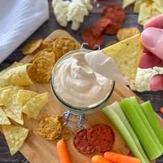 a person holding a tortilla chip over a bowl of dip surrounded by veggies and crackers