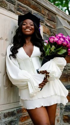 a woman in a white dress holding flowers and smiling at the camera while leaning against a brick wall