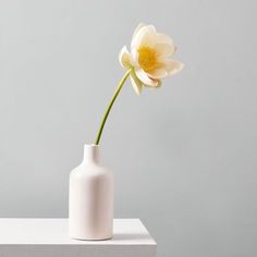 a white vase with a yellow flower in it on top of a table next to a gray wall