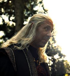 a man with long blonde hair wearing a black outfit and standing in front of trees