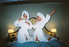 two women in white robes sitting on a bed with their arms up and one holding a cup