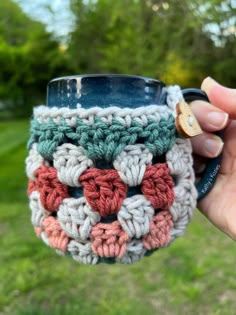 a hand holding a crocheted coffee cup in front of a grassy area with trees