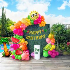 a happy birthday sign surrounded by balloons and fruit in front of a pool with palm trees