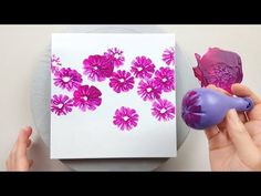 two hands are holding purple and pink flowers in front of a white paper on a gray plate