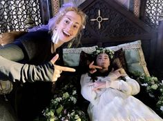 two women dressed in medieval garb lying on a bed with flowers and greenery