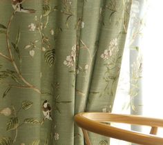 a chair sitting next to a window covered in green floral curtain panels with birds on them