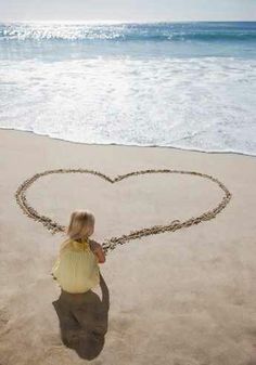 a woman standing on top of a beach next to the ocean drawing a heart in the sand