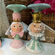 two ceramic figurines sitting on top of a table next to pink flowers and baskets