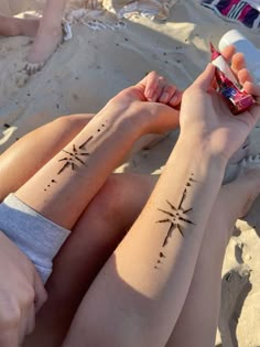 two people with matching tattoos on their arms sitting in the sand and one is holding a cell phone