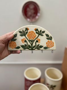 a hand holding up a decorative object with orange and green flowers on it, in front of several coffee cups