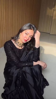 a woman sitting on the ground wearing a black dress and necklace with her hands in her hair