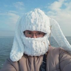 a person wearing a knitted white bunny hat and scarf over their face with the ocean in the background