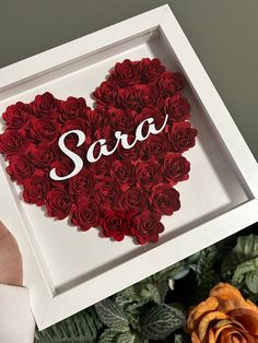 a box with roses in the shape of a heart that says sara on it and is surrounded by greenery