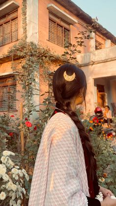 a woman standing in front of a building with flowers and vines around her head, looking down at the ground