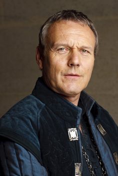 a man with grey hair wearing a black vest and chain necklaces, looking at the camera