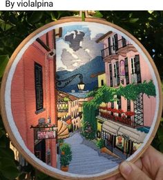 a hand embroidered picture of a street with buildings and trees in the background, on a wooden frame