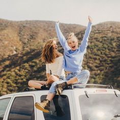 two women sitting on the roof of a white car with their arms in the air