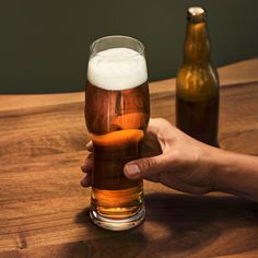 a person holding a beer in their hand on a wooden table with two bottles behind them