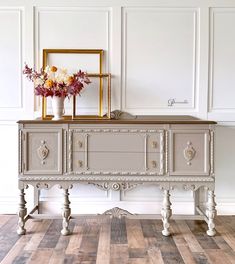 an old dresser with flowers on top in a white room next to a gold frame